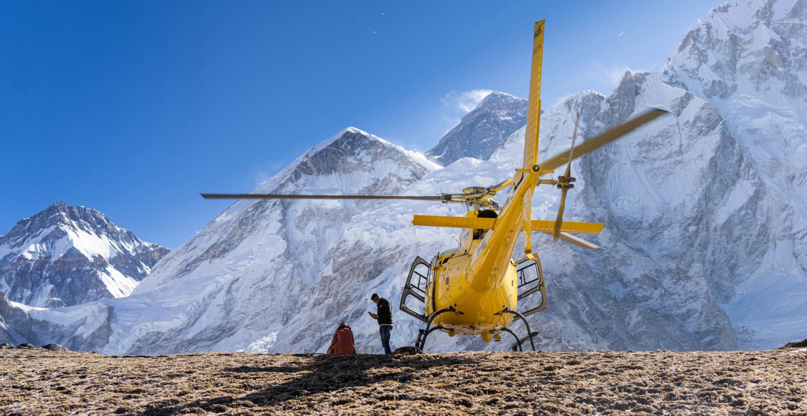 A bright yellow rescue helicopter sits on a rocky, brown plateau with its rotors spinning.